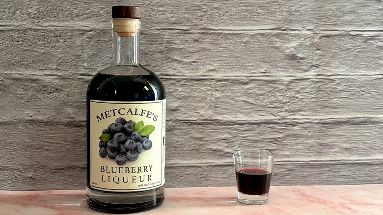 Bottle of Metcalfe's Blueberry Liqueur  next to shot glass of liqueur on grey brick background