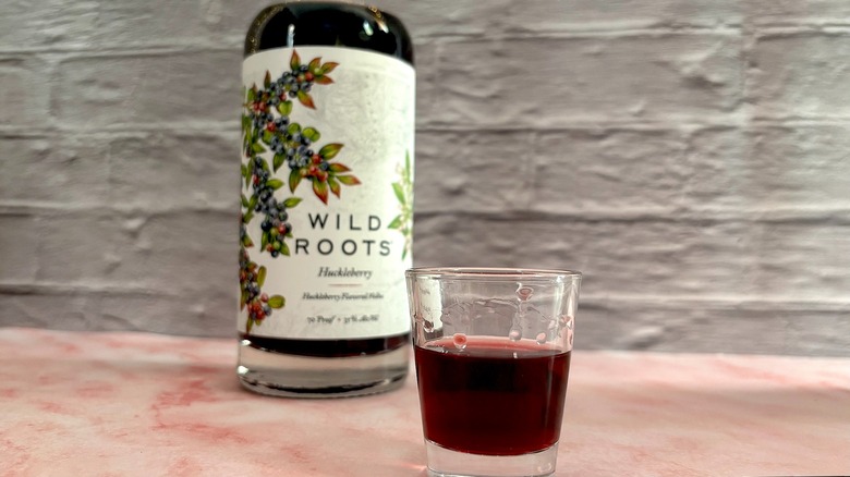 Bottle of Wild Roots Huckleberry Vodka behind shot glass of vodka in front of grey brick wall