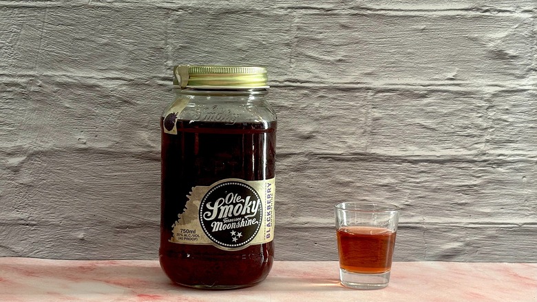 Jar of Ole Smoky Blackberry Moonshine  next to shot glass of moonshine on grey brick background