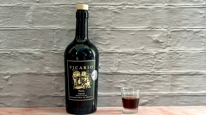 Bottle of Vicario Mirto Liqueur  next to shot glass of liqueur on grey brick background