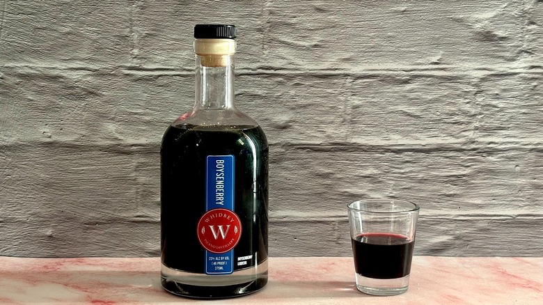 Bottle of Whidbey Island Distillery Boysenberry Liqueur next to shot glass of liqueur on grey brick background