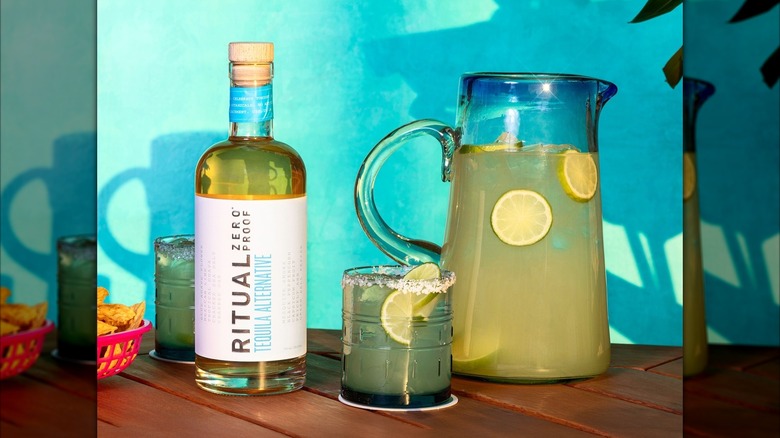 Bottle of Ritual tequila alternative next to glass and pitcher of lemonade on wooden table, blue wall background