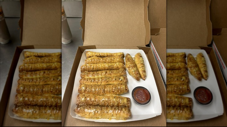 Garlic Parmesan breadsticks in cardboard takeout box with pot of marinara sauce