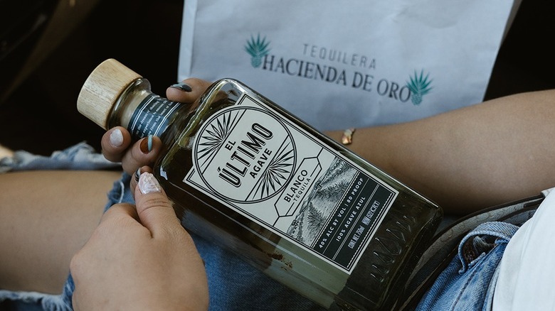 A woman holding a bottle of El Último Agave Blanco with a bag in the background