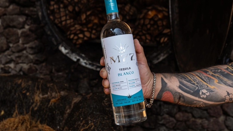 A tattooed arm holding a bottle of Lote M77 Blanco tequila