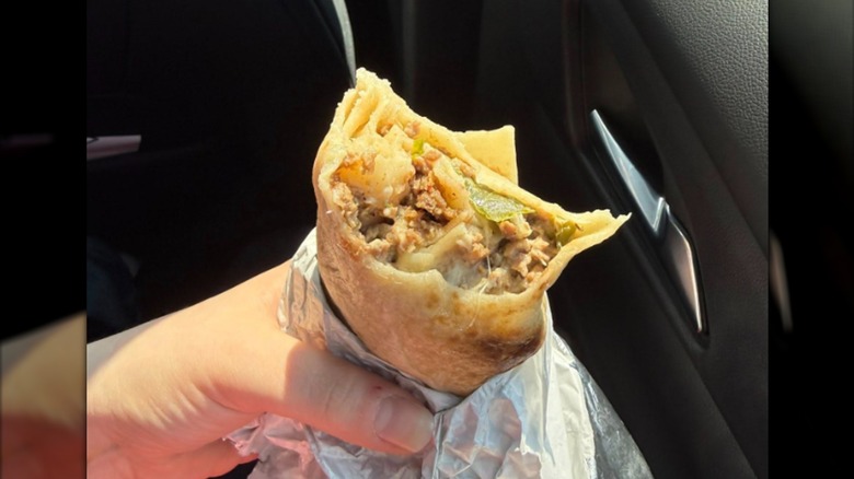 A hand holding a half-eaten Texas Cheesesteak Burrito partially wrapped in foil inside a car