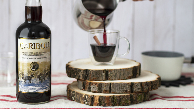 Bottle of Caribou liquor next to stacks of log rounds with cup of Caribou and hand pouring liquid from a pot