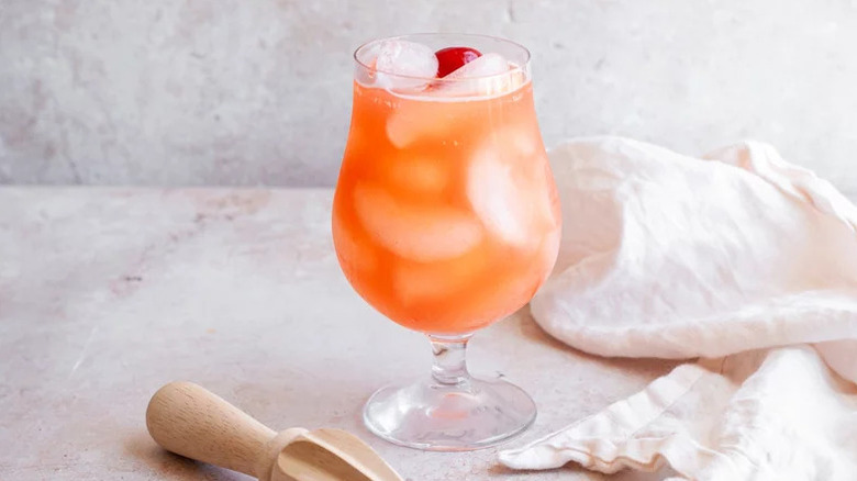 Coral pink drink over ice in a snifter against textured white background with towel and juicer.