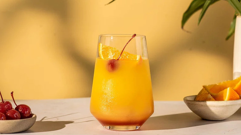 A Tequila Sunrise in a glass with orange and cherry garnish on marble table next to bowls of fruit.
