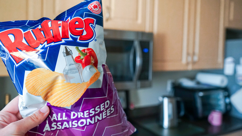 Bag of Ruffles chips with the flavor of "all dressed" held up by a hand with a kitchen in the background