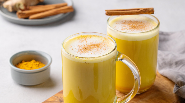 golden milk in clear mugs with cinnamon sticks and jar of turmeric powder