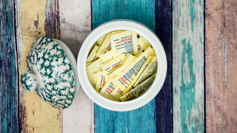 An opened jar filled with artificial sweetener packets sits on a colorful wooden plank background