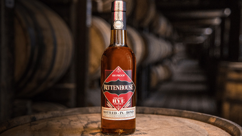 A bottle of Rittenhouse Rye on top of a barrel in a warehouse with other barrels
