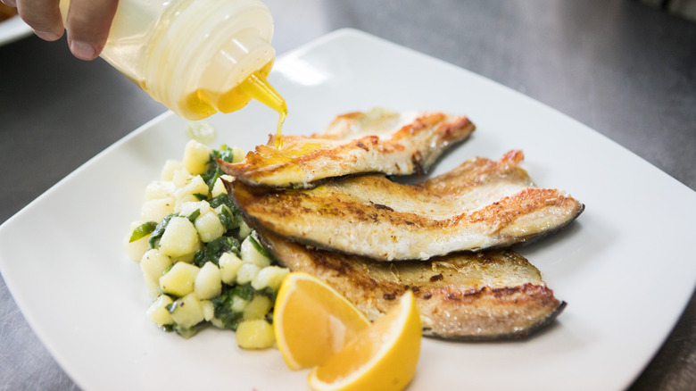 hand pouring oil from bottle on to roasted trout with side veg and lemon on white dish