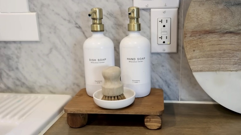 Small wooden tray holding white soap bottles and saucer with scrub brush inside