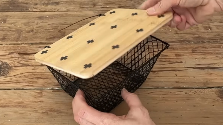 Hands lifting wooden lid of small black wire basket sitting on wood surface