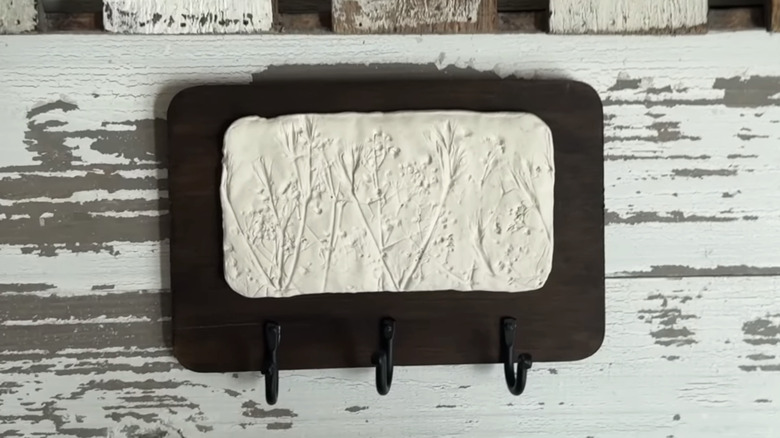 Dark wood board with three black hooks and decorative floral clay panel hung on worn white wall