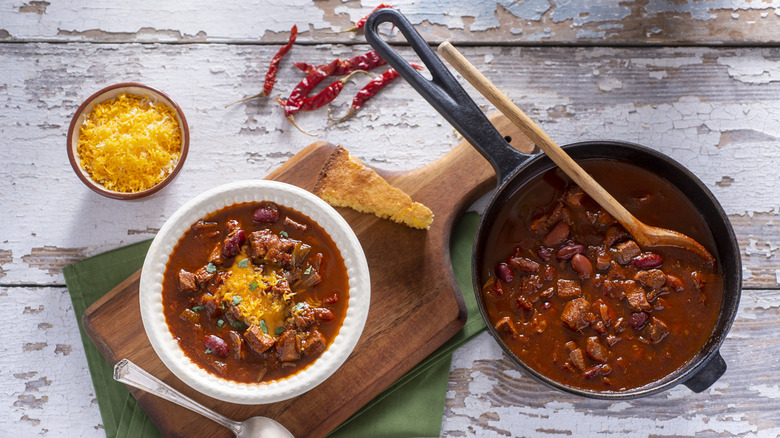 Bowl of chili con carne on cutting board with cheese and cornbread