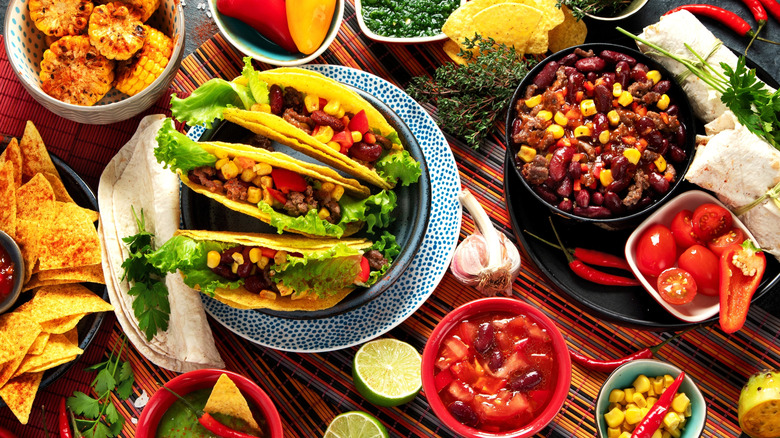 Hard shell tacos surrounded by corn, tomatoes, tortilla chips, and chili