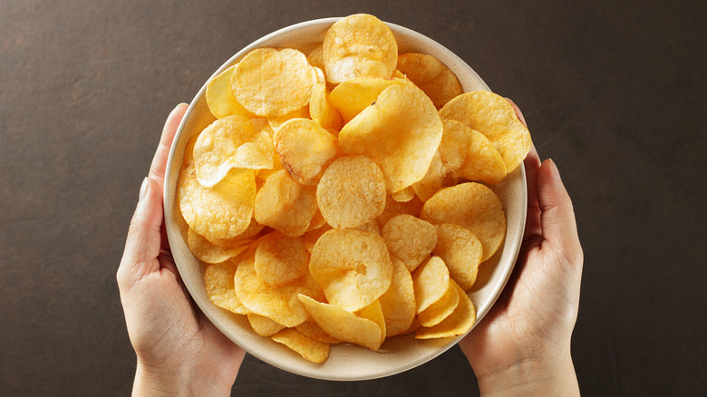Hands around bowl of potato chips on dark brown surface