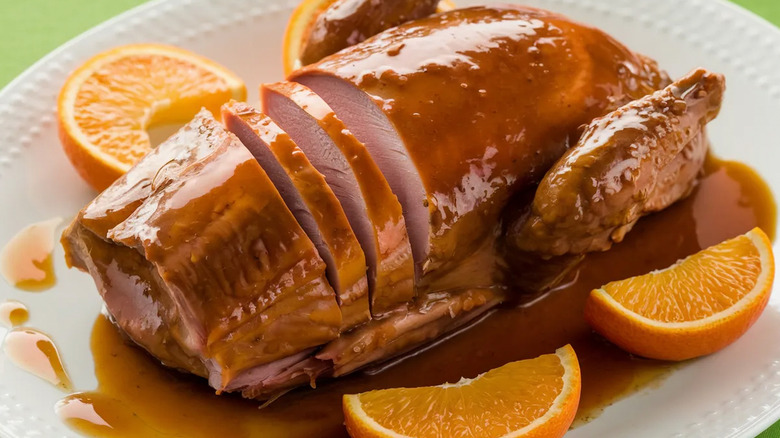 Whole roasted duck glazed in glossy orange sauce with orange slices on white plate.