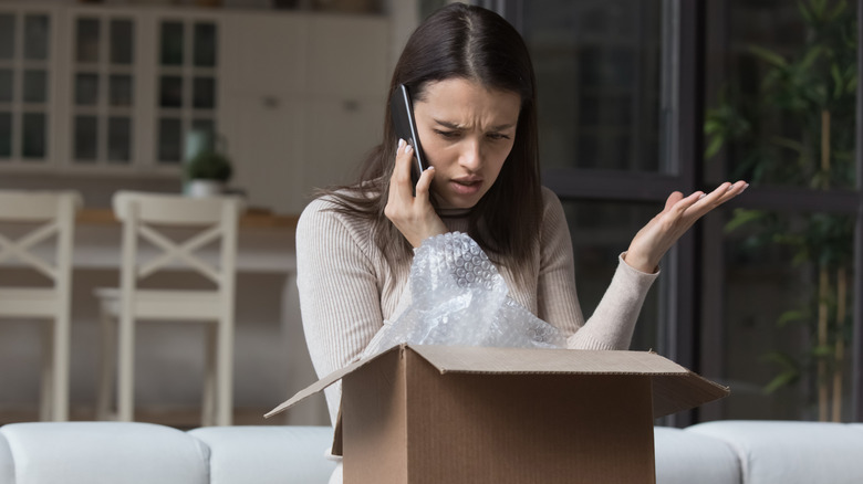 Annoyed-looking woman in a pale beige sweater, looking into a newly-opened box, and holding her phone to her ear