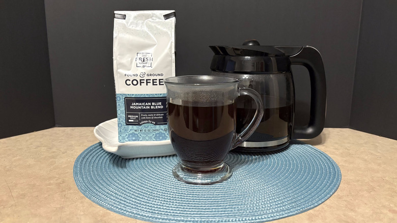 The Fresh Market Jamaican Blue Mountain blend coffee in bag, pot, and glass on teal placemat and black background