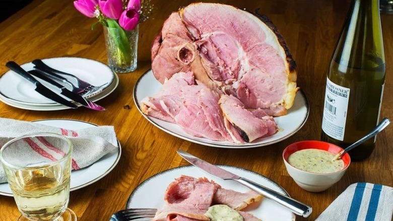 Baked ham served onto plates next to bottle of wine and bowl of creamy sauce