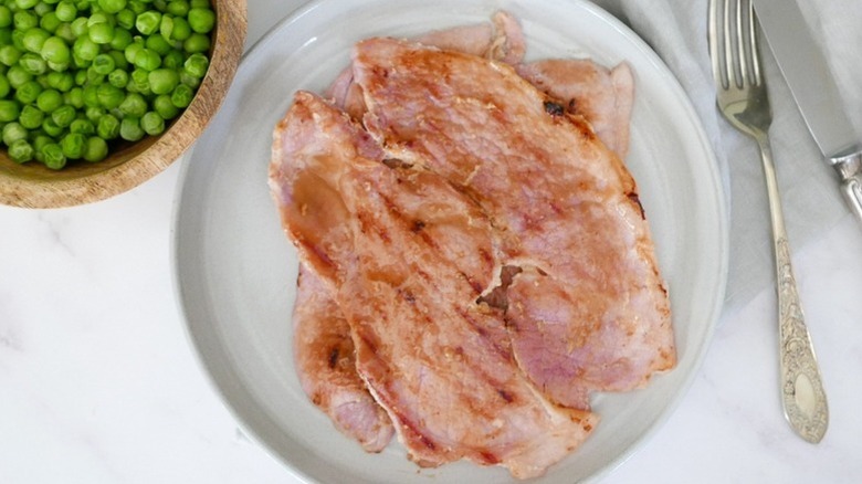 Sliced of glazed ham on plate next to bowl of peas
