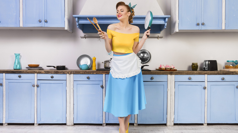 Vintage retro-styled 1960s era woman in light blue kitchen holding frying pan and wooden utensils
