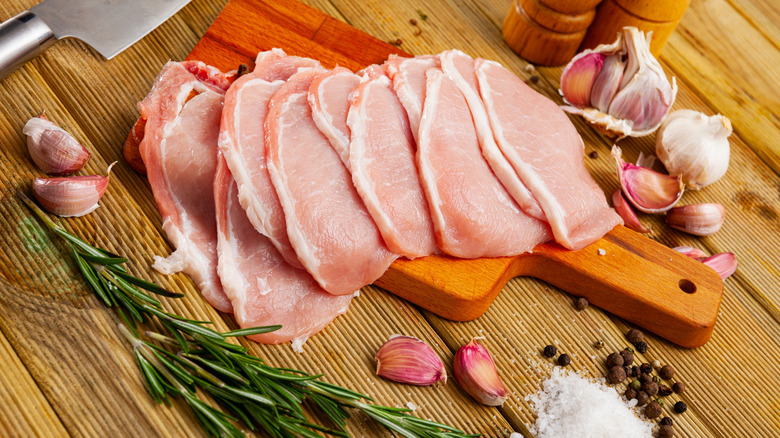 a stack of thin pork loin chops on a wooden cutting board on a rustic wooden work surface, surrounded by garlic cloves, fresh rosemary and other seasonings