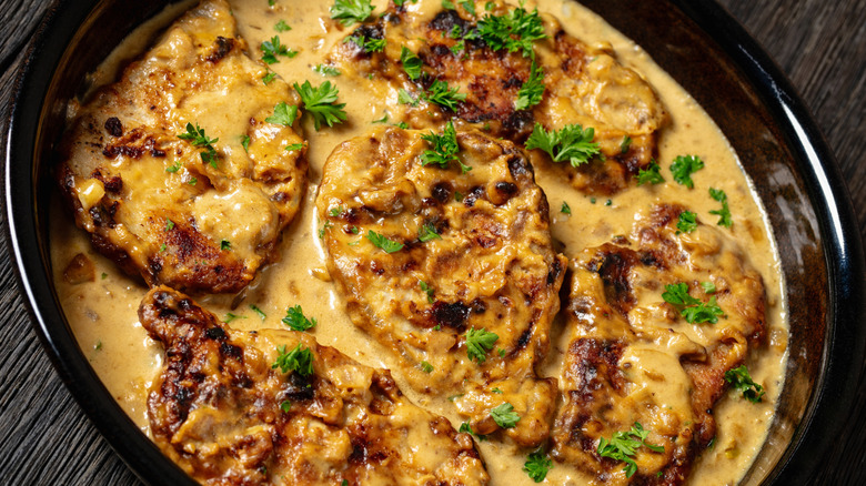 smothered pork chops in a creamy sauce, shown in a dark-colored baking dish on a rustic wooden surface