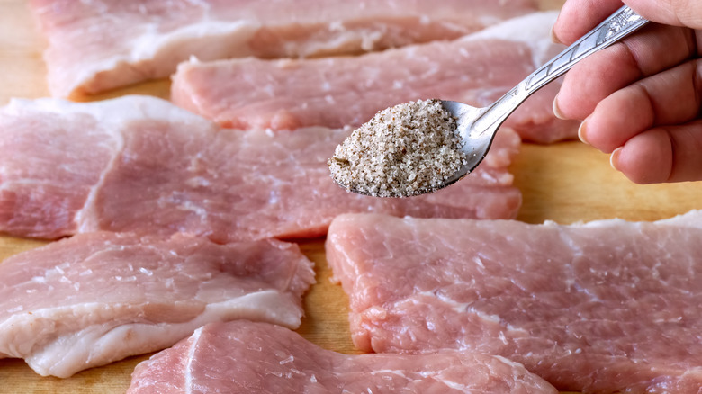 female hand sprinkling seasonings from a spoon onto uncooked pork chops