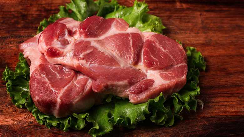 two pieces of raw pork leg steak on a frilly lettuce leaf on a dark wood surface, showing the complicated muscle structure of the cut