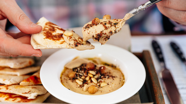 A hand scoops hummus with nuts and seeds onto a slice of pita