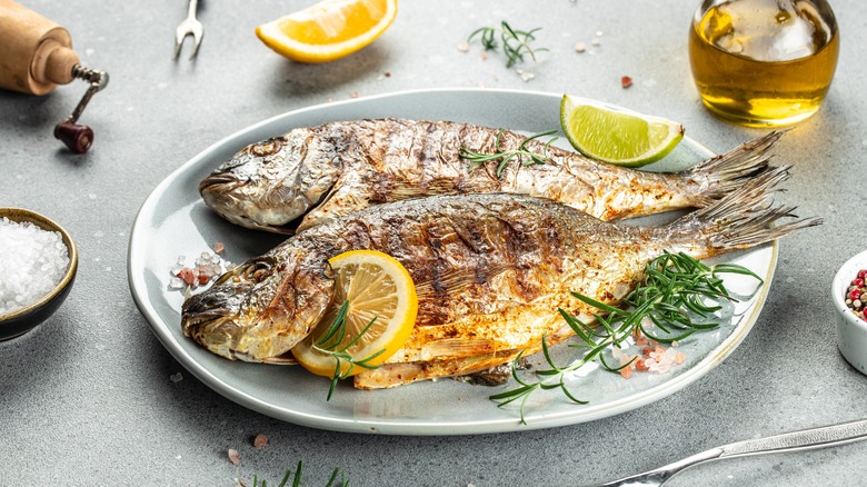 Two whole grilled fishes on a white plate with lemon and herbs