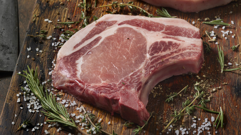 bone-in pork rib chop on a cutting board, surrounded by peppercorns, coarse salt, and fresh rosemary and thyme