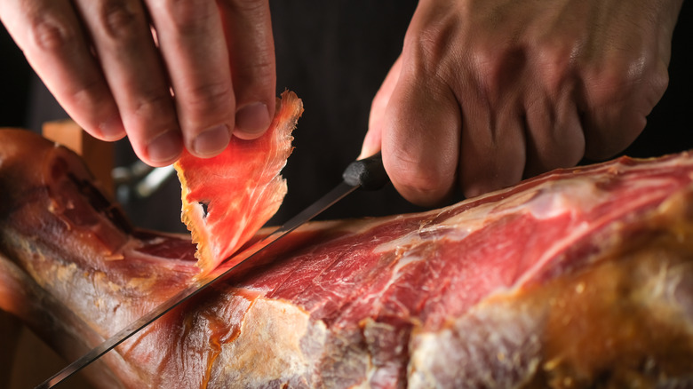 man's hands slicing a thin shaving from a whole dry-cured ham, perhaps Italian prosciutto or Spanish jamon