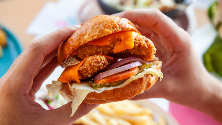Hands holding a fried chicken sandwich with cheese, lettuce, tomato, and onion