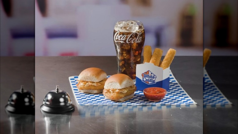 Two White Castle Garlic Parm Chicken Slider sandwiches on blue and white checkered placemat next to cup of Coca Cola, sleeve of mozzarella sticks, and cup of red sauce