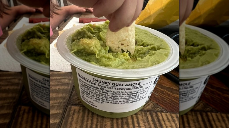 Hand dipping chip into open pot of chunky guacamole with busy family table in the background