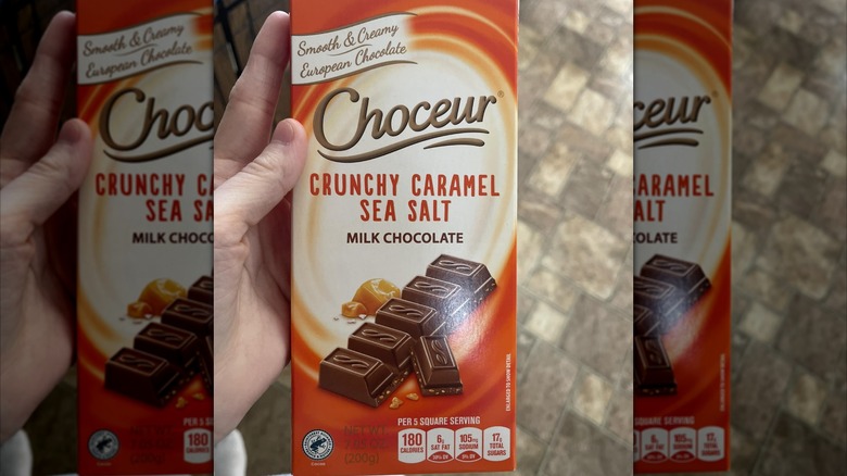 Hand holding bar of Choceur Crunchy Caramel Sea Salt over brown floor tiles, mirrored image