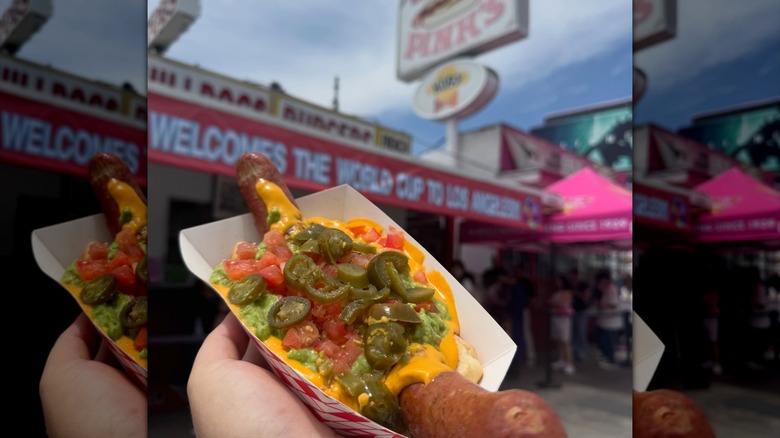Hand holding loaded hot dog in tray in front of blurred Pink's building