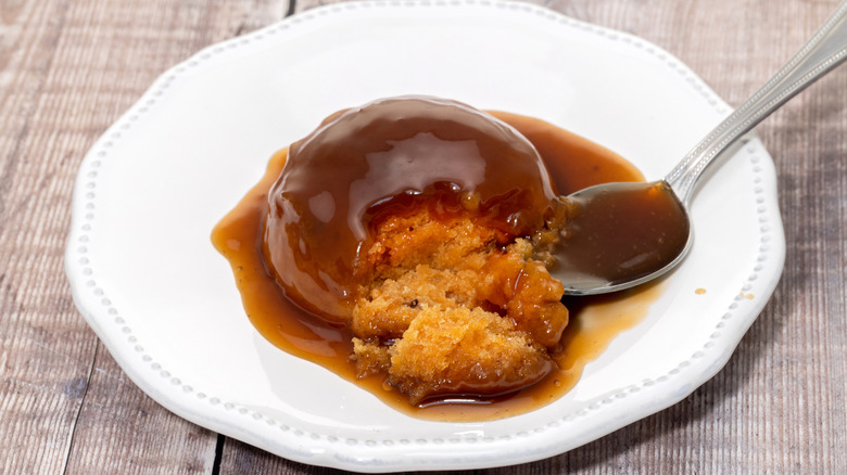 A sticky toffee pudding on a white plate with spoon on a wooden table