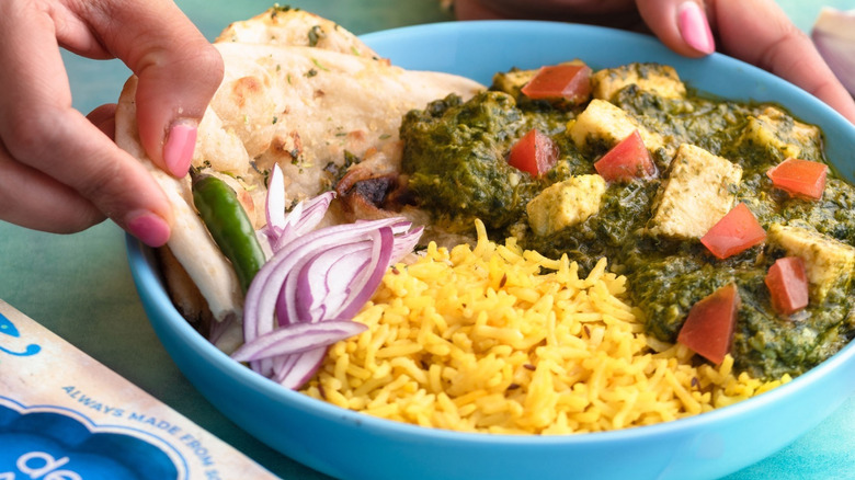 A bowl of spicy paneer with rice, garlic naan, onions, chilies, on a blue table