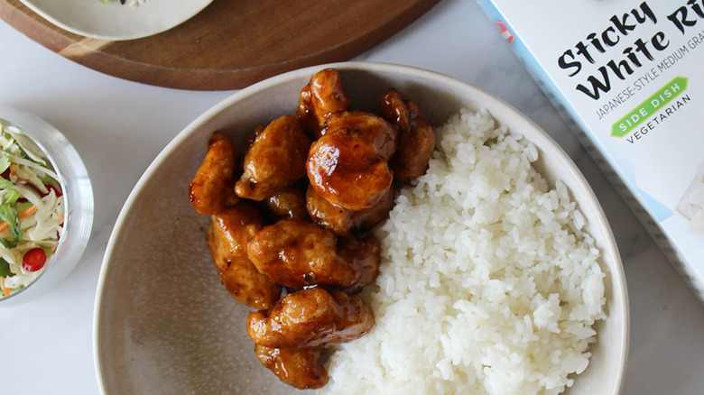 A plate of InnovAsian general tso's chicken and sticky white rice on a white countertop surrounded by brand boxes, vegetables, and fried egg