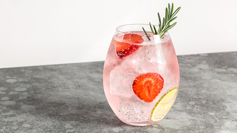 A glass of pink gin on a countertop with ice, strawberries, rosemary, and lime added.