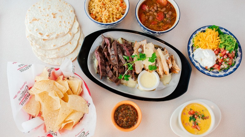Steak and chicken fajita meat on platter with tortillas, chips, cheese, rice, beans, and toppings in side bowls