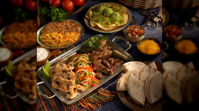 Tray of fajita steak and chicken with veggies and side plates with tortillas, rice, beans, and guac