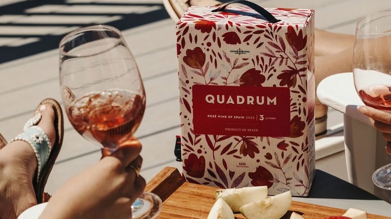 Box of wine beside a person holding wine glass with fruit on table